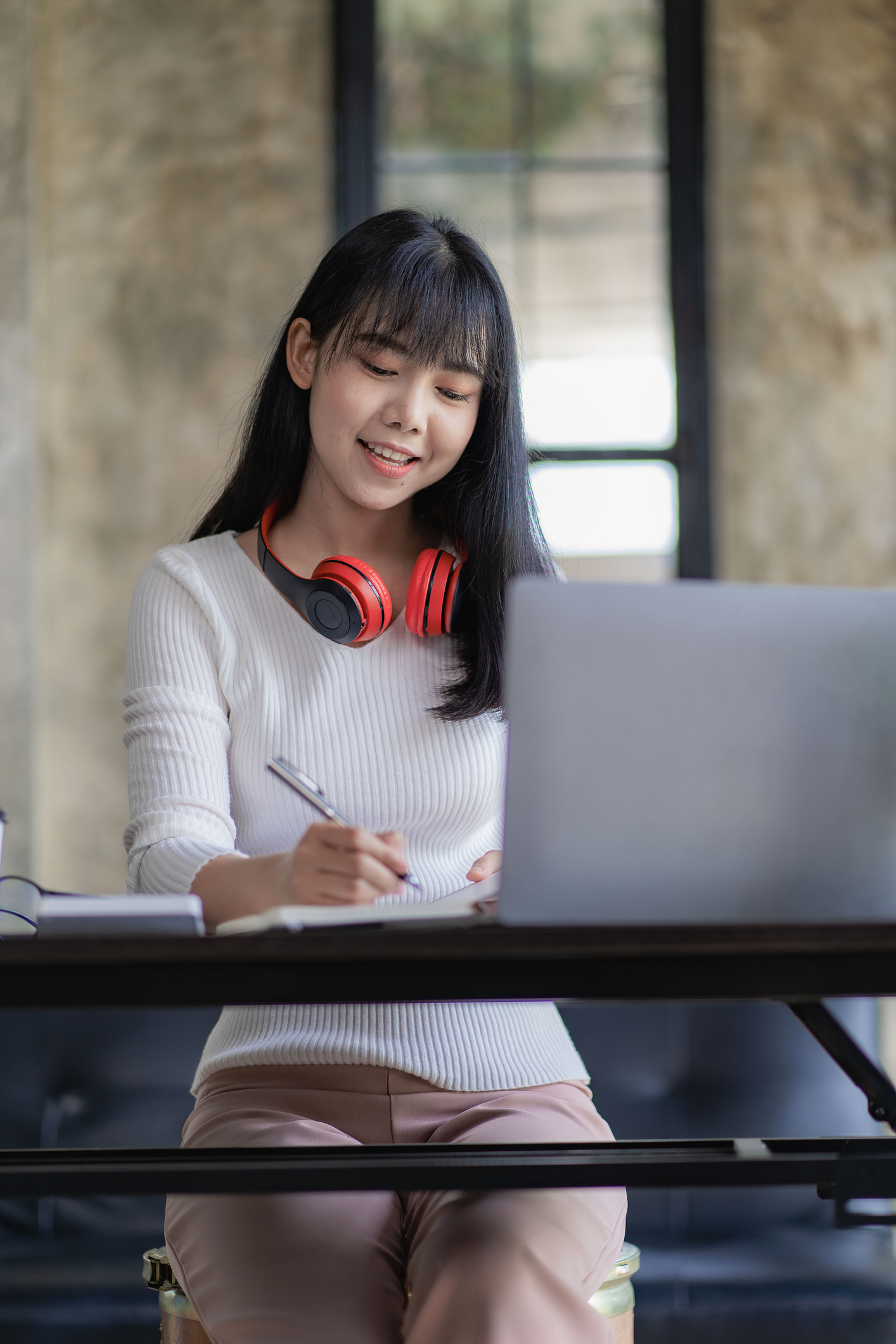 Beautiful and cheerful Asian woman wearing headphones in video conferences and online classes on the Internet and enjoying listening to music happily on her work laptop during work breaks.